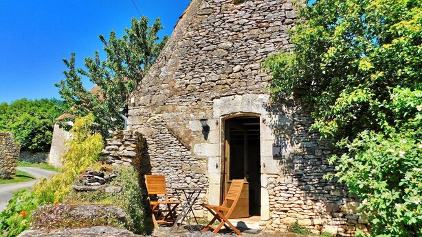 Une petite maison en pierre avec une porte en bois, une chaise à l’extérieur et de la verdure tout autour.