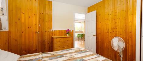 Een slaapkamer met houten wanden, een bed met sprei, een dressoir, een ventilator en een deur naar een andere kamer.