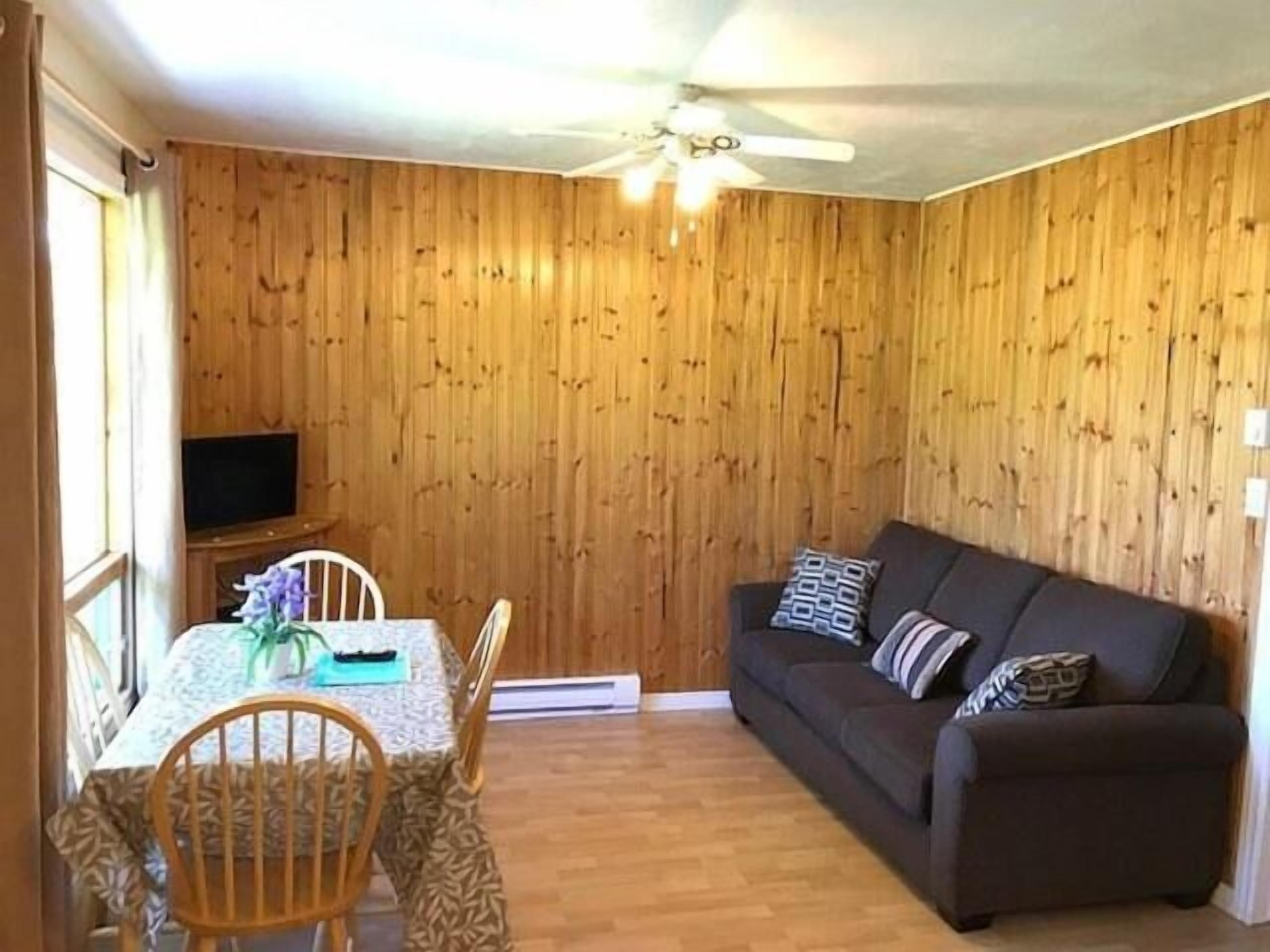 A room with a wooden paneled wall, a ceiling fan, a sofa with patterned cushions, a dining table with chairs, and a television on a stand.