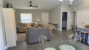 A spacious living room with a grey sofa, a round glass table with a decorative centerpiece, a dining area with a chandelier, and a kitchen in the background. - New modern 3-bed 2 bath house with smart TV's in every room, WIFI, and hot tub (Fort Pierce)