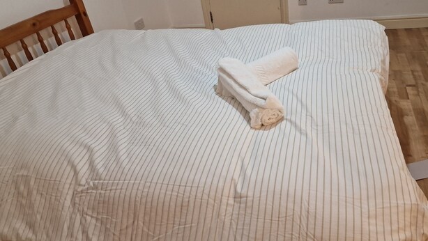 A bed with a striped white and gray bedspread, a wooden headboard, and a rolled white towel on the bed. - Retreat Studio Camberwell zone 2 (London)