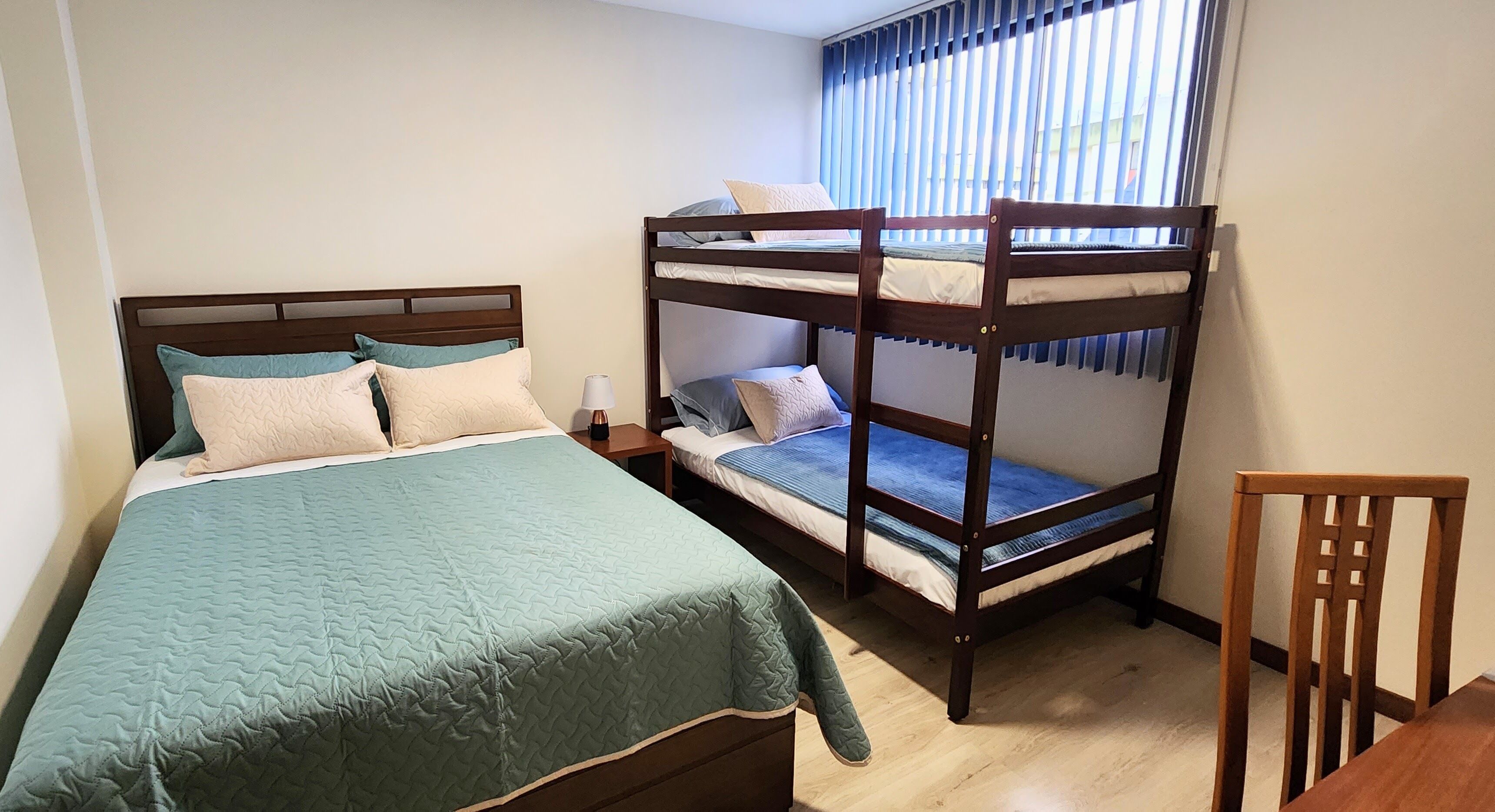 Une chambre avec un lit superposé, un lit simple, une chaise en bois et une table de chevet avec une lampe.