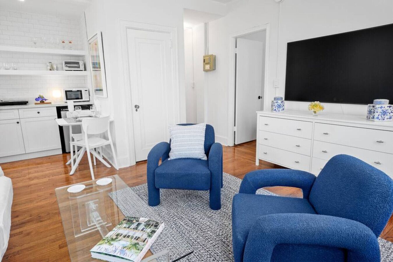 A modern living room with a blue sofa, a coffee table, and a TV. There is a kitchen area with white cabinets and a microwave.