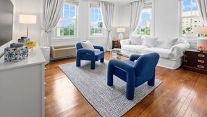 A modern living room with a white sofa, two blue armchairs, a coffee table, and a rug. There are two windows with curtains, a ceiling light, and a white cabinet with decorative items.