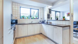 Une cuisine moderne avec des meubles blancs, un four encastré et une machine à café. De grandes fenêtres offrent une vue sur la verdure environnante.