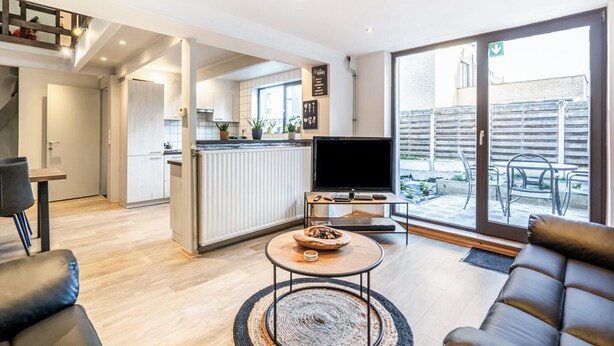 Un salon moderne avec un téléviseur à écran plat, un canapé et une table basse. Il y a également un coin cuisine équipé d’un réfrigérateur et d’une table à manger. La pièce est dotée de grandes fenêtres et offre une vue sur l’extérieur.