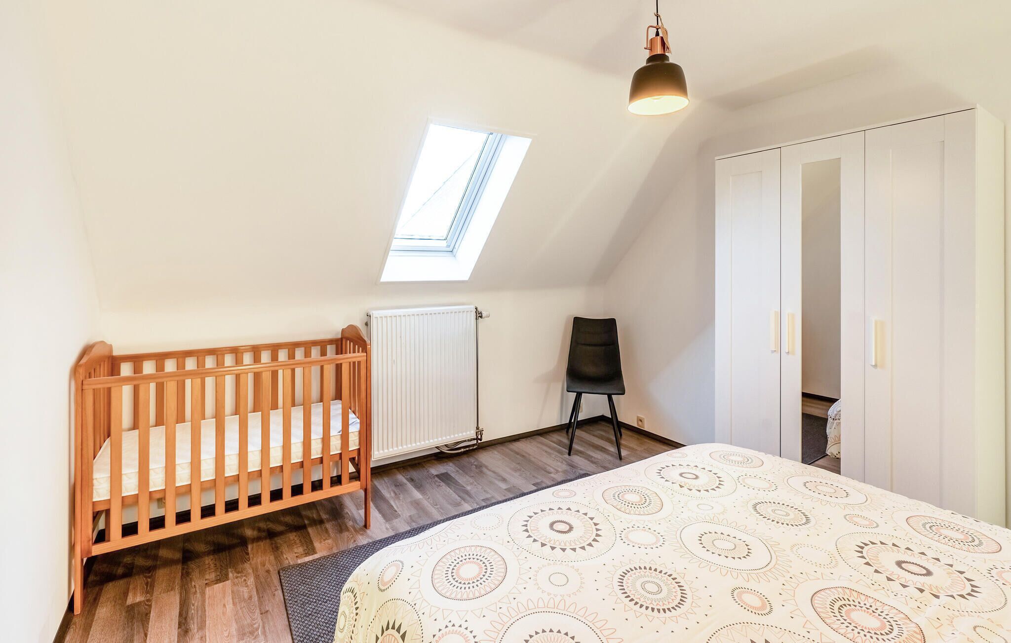 A room with a sloped ceiling, a wooden crib, a patterned bedspread, a radiator, a chair, and a wardrobe.