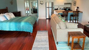 A compact living space with a bed, sofa, wooden floor, kitchen area, and a ceiling fan. - Spacious Studio Near Beaches & Dining (Waikoloa)