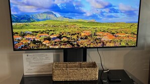 A flat-screen TV mounted on a wooden cabinet, displaying a scenic landscape with mountains and a village. - Spacious Studio Near Beaches & Dining (Waikoloa)