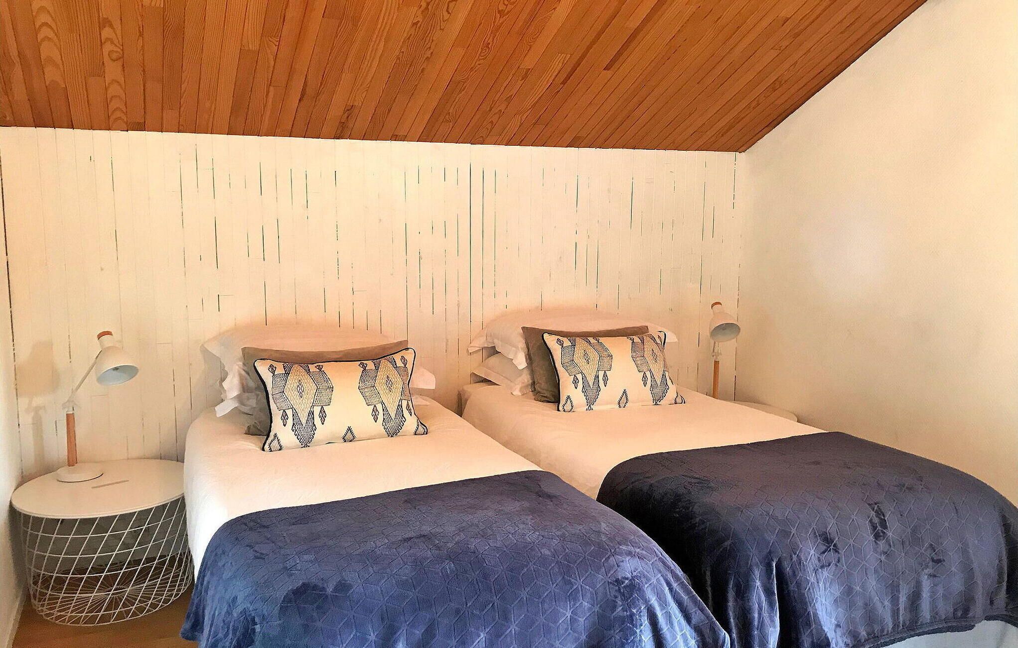Une chambre avec deux lits, chacun agrémenté d’oreillers à motifs et d’une couvre-lit bleue. On y trouve des lampes de chevet et une petite table d’appoint ronde, dont le cadre est en fil de fer.