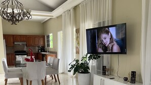A spacious living room with a dining area, a kitchen in the background, and a large flat-screen TV mounted on the wall. - Casa de la Playa (Puerto Plata)