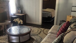 A cozy living room with a round coffee table, a sofa with patterned cushions, and a view into a bedroom through an open door. - Cute cottage in a rural setting (Clemmons)