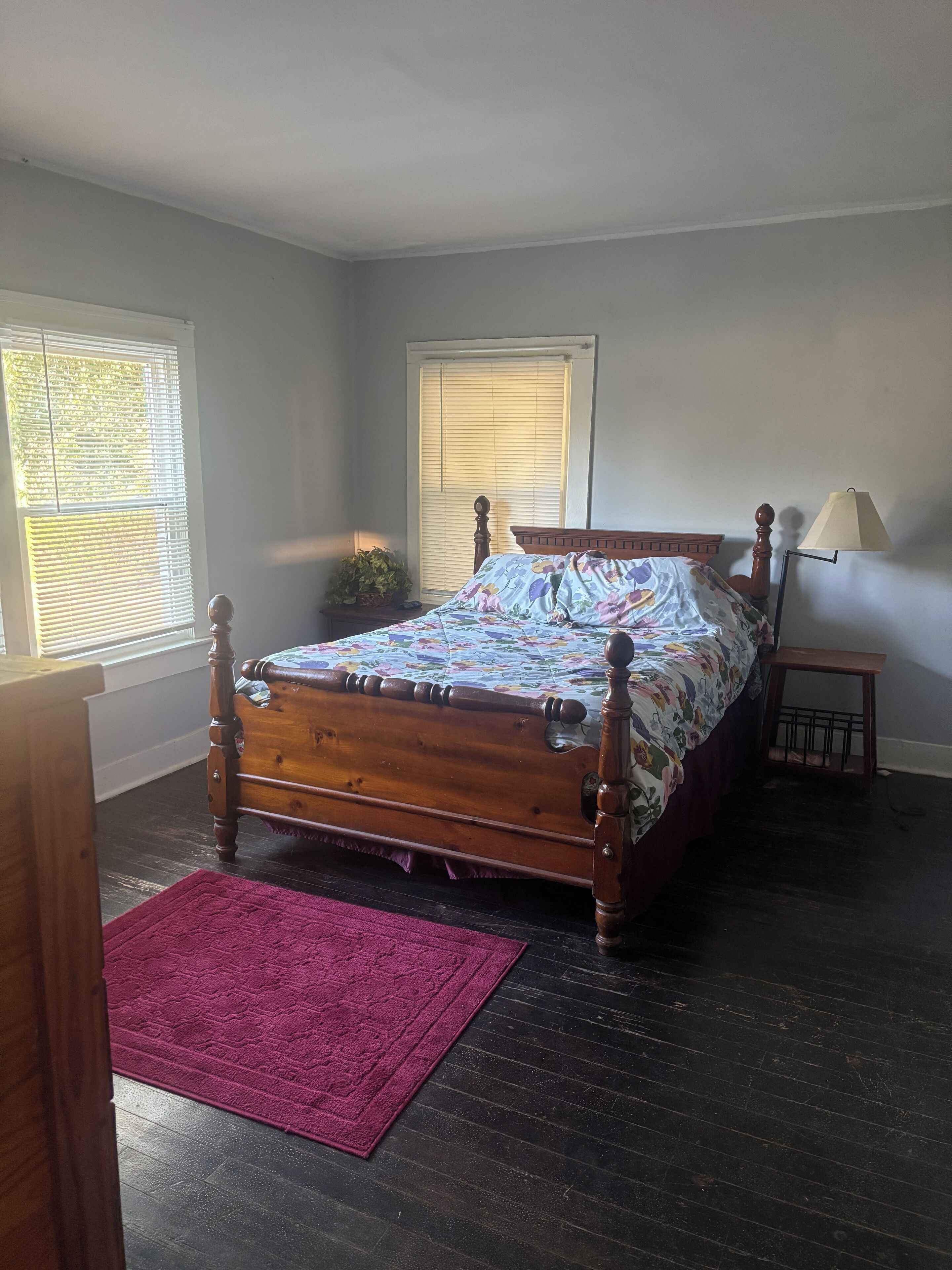 Een slaapkamer met een houten bed, een rode vloerkleed, een nachtkastje, een lamp en een raam met jaloezieën.