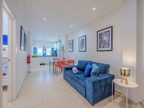 A modern living room with a blue sofa, a small side table, and a dining area with red chairs.