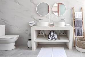 A modern bathroom with a white toilet, two round mirrors, a white sink with a marble countertop, and a wooden towel rack with towels. - Belvilla Serene Haven Nusa Dua Beach (Nusa Dua)