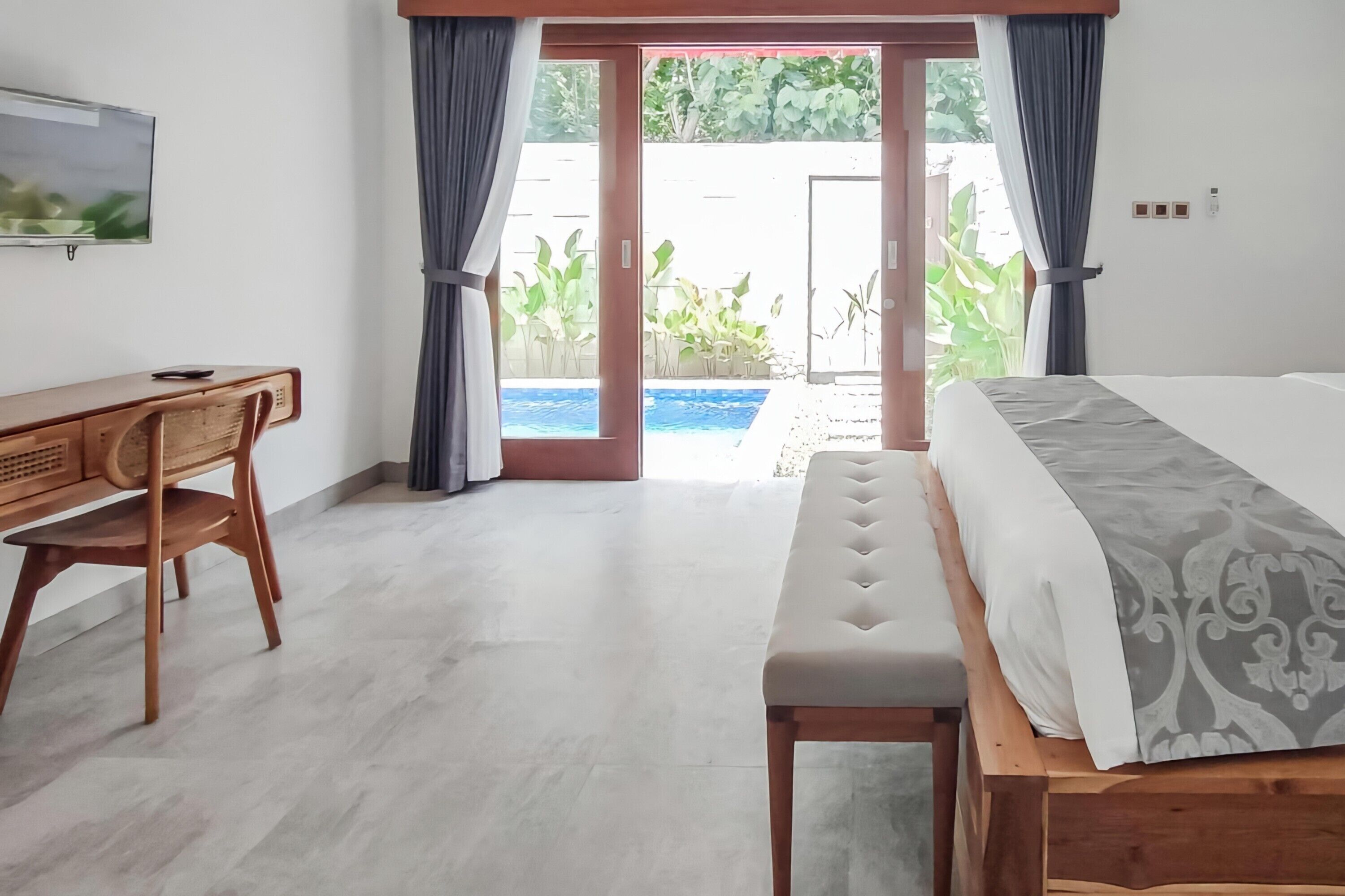 A bedroom with a bed, a desk, and a bench. There is a sliding glass door leading to a balcony with plants.