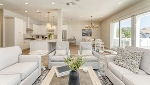 Una sala moderna con sofá, sillones y una mesa de centro. Hay una zona de cocina con armarios blancos y una mesa de comedor.
