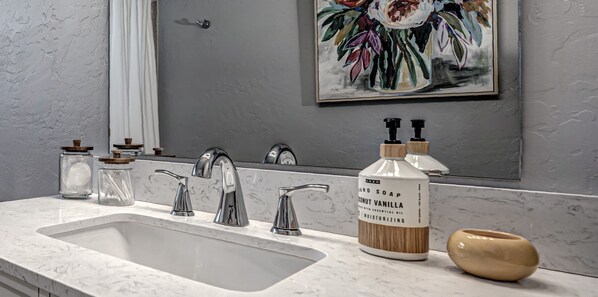 A bathroom with a marble sink, three faucets, a large mirror, a floral painting, and a bottle of coconut vanilla hand soap. - Bright 3BR 2BA Entire Home in Norman-Close to OU! (Norman)
