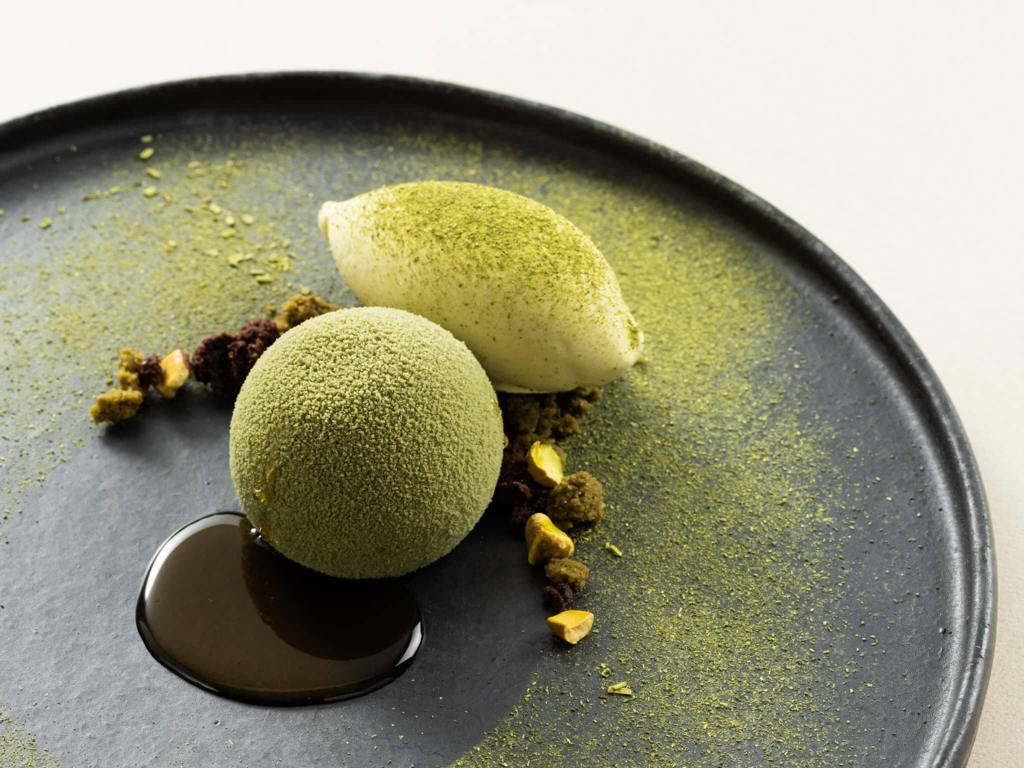A dessert with two spherical sweets on a dark plate, accompanied by crushed nuts and a small pool of syrup.