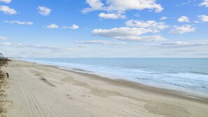 A wide sandy beach with gentle waves, a clear sky, and a distant shoreline with buildings.