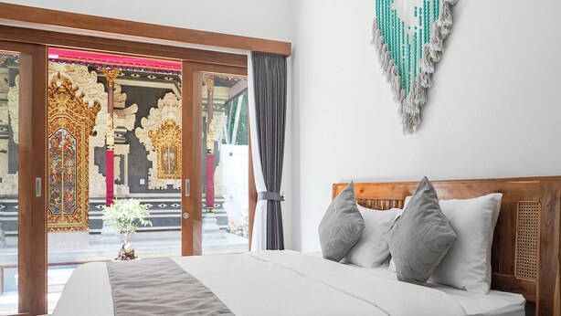 A hotel room with a large bed, wooden headboard, and a view of traditional architecture through a glass door. - Belvilla Calm Woods Pandawa Beach (Nusa Dua)