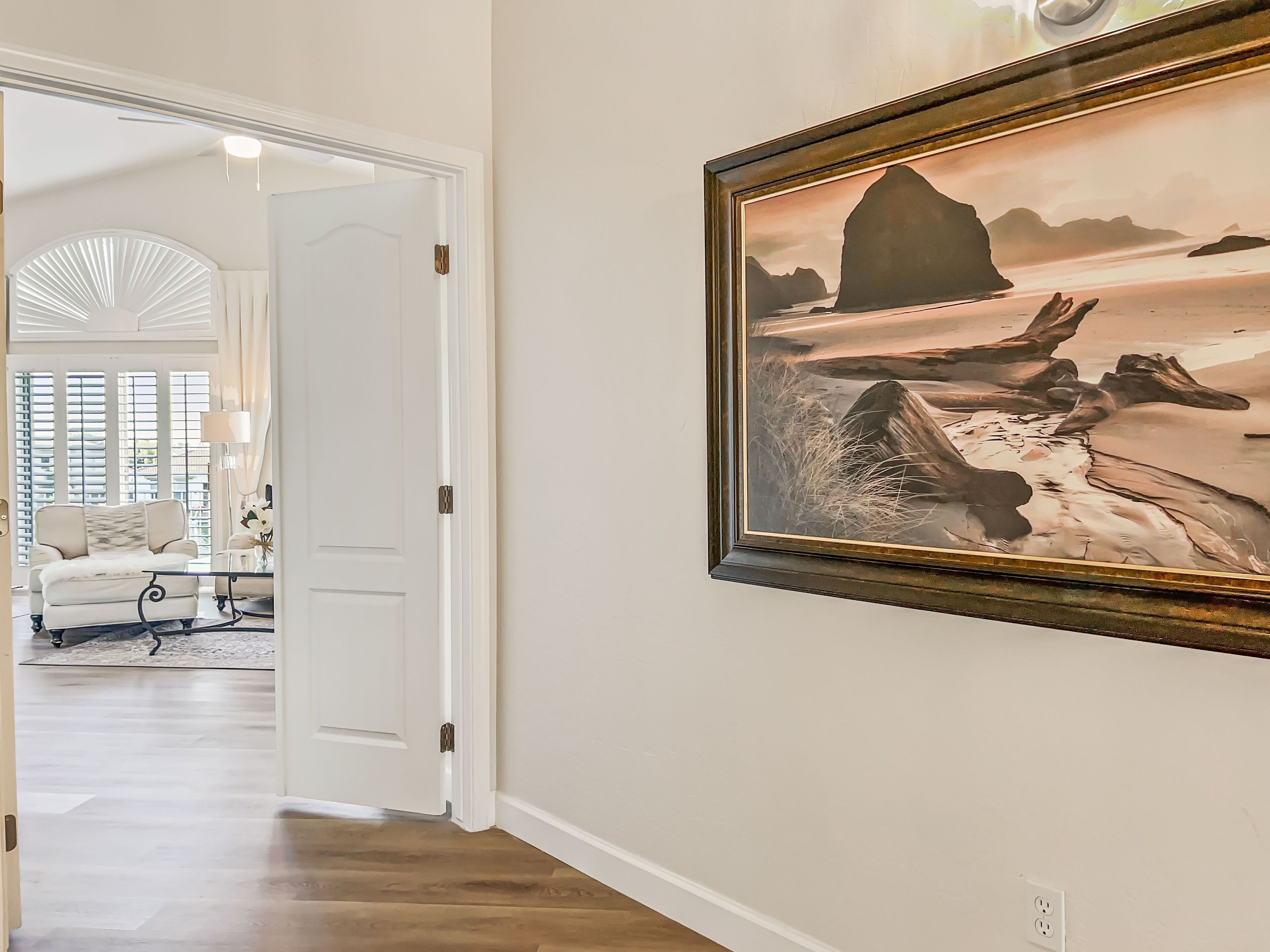 A hallway with a framed landscape painting, a white door, and a glimpse of a living room with a sofa and a lamp.