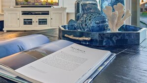 A living room with a television, decorative items, and a book on a table. - 5BR w lake view, heated pool, hot tub, sauna (Gilbert)