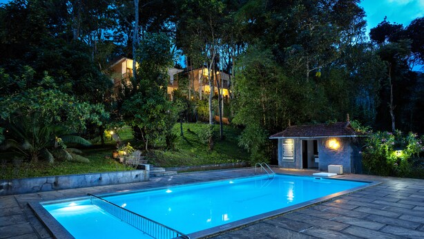 A swimming pool with a stone deck, surrounded by lush greenery and a small pavilion. - Coffee Routes Thekkady (Peermade)