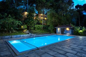 A swimming pool with a stone deck, surrounded by lush greenery and a small pavilion. - Coffee Routes Thekkady (Peermade)