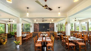 A spacious dining area with multiple tables and chairs, ceiling fans, and large windows offering views of greenery. - Coffee Routes Thekkady (Peermade)