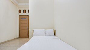 Un couloir étroit mène à une porte sur laquelle est apposée une pancarte. Un lit recouvert de draps blancs est placé contre le mur.