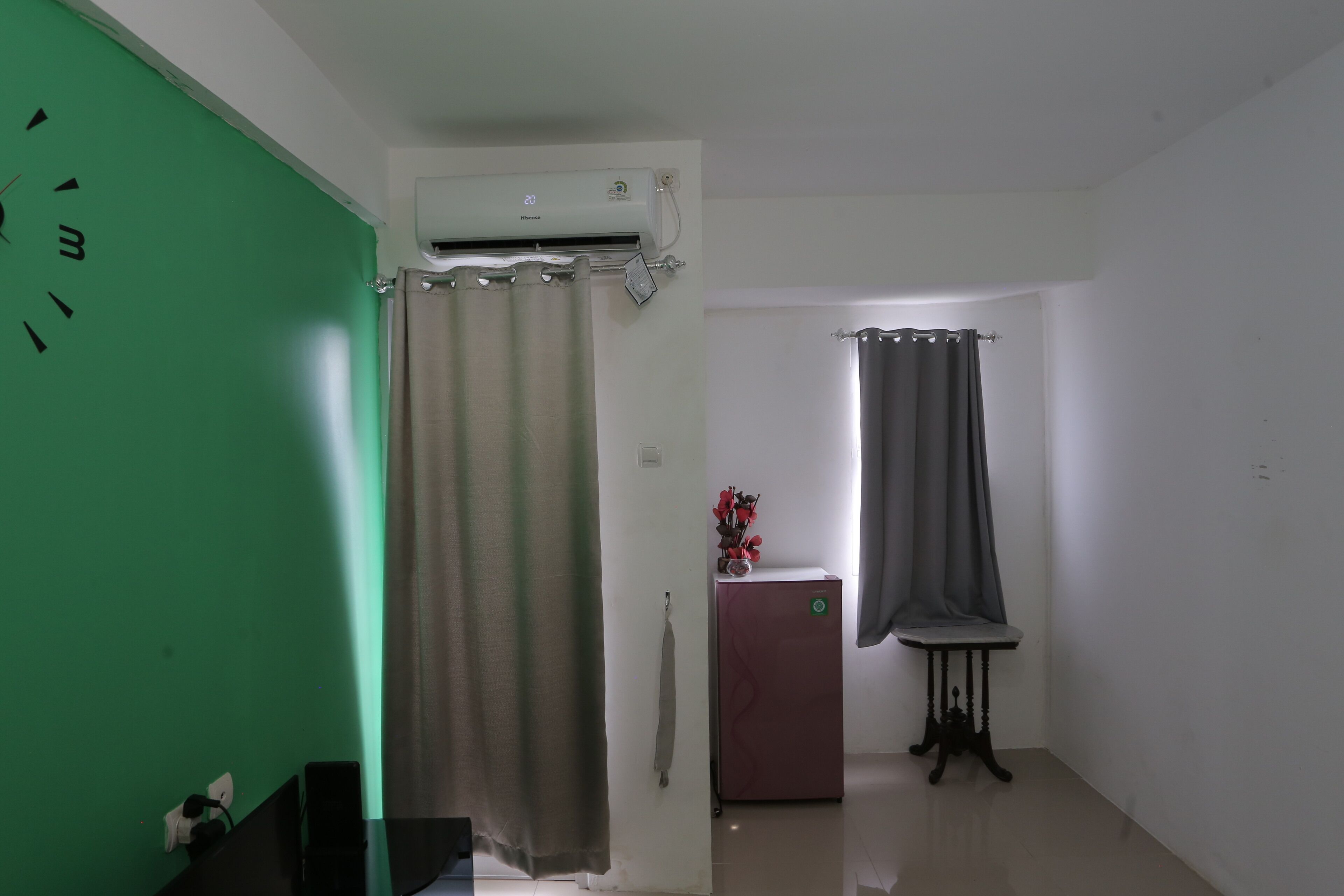 A room with a green wall, a beige curtain, a small refrigerator, a table with a vase, and a window with a grey curtain.