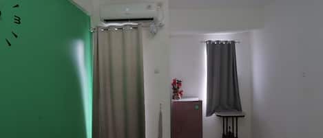 A room with a green wall, a beige curtain, a small refrigerator, a table with a vase, and a window with a grey curtain.