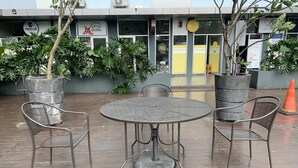 Ein Sitzbereich im Freien mit einem runden Tisch und vier Stühlen, umgeben von Topfpflanzen und einem Gebäude mit verschiedenen Werbeschildern im Hintergrund.