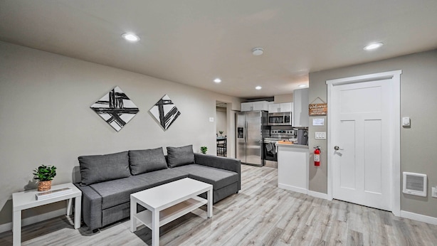 A modern living room with a grey sectional sofa, a white coffee table, and a small side table with a plant. There is a kitchen area in the background with a refrigerator, microwave, and stove. - Nicely remodeled 3 bed house in Seattle - Free parking (Seattle)