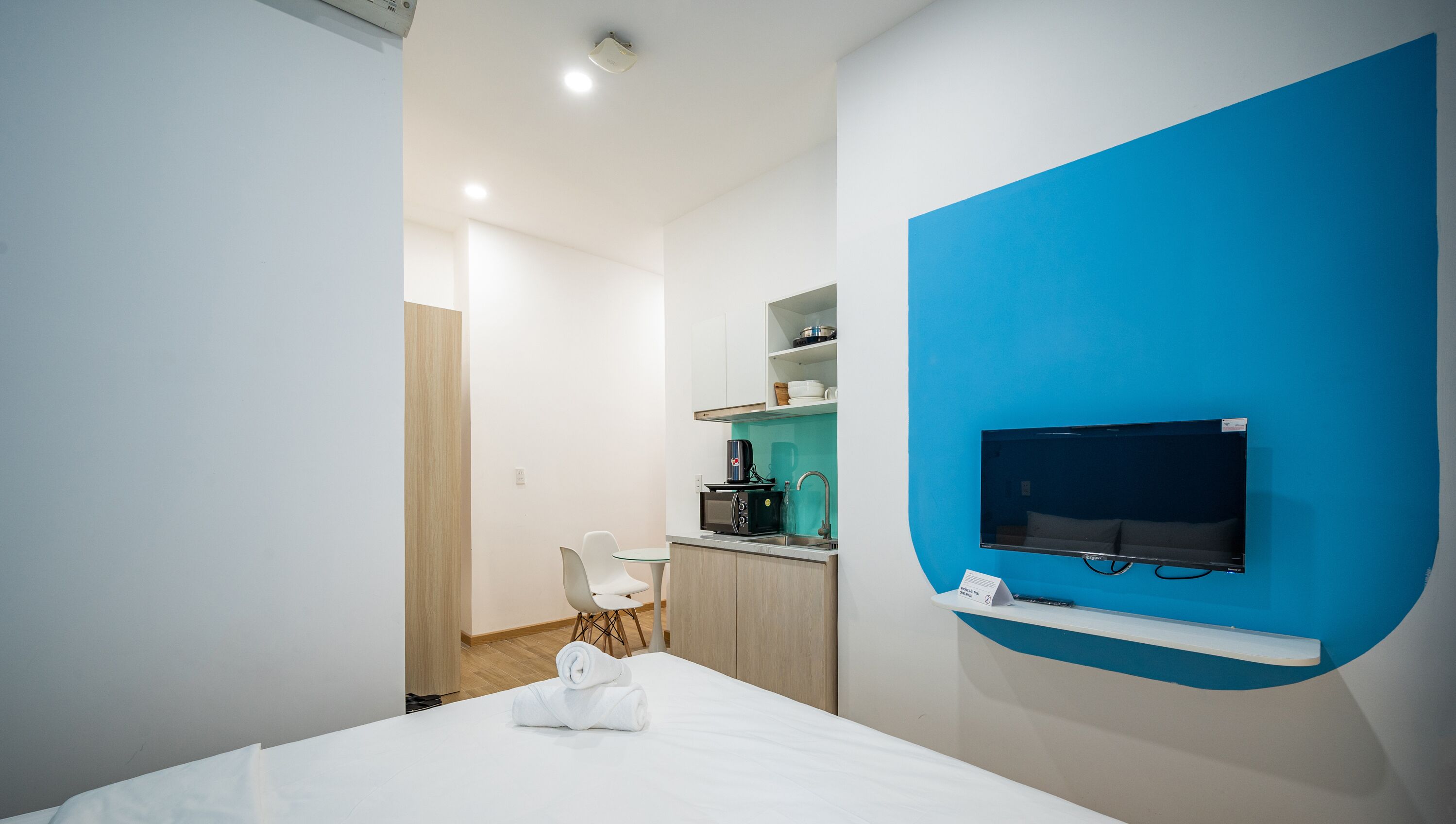 A modern hotel room with a bed, a TV mounted on a blue wall, a kitchenette with a coffee machine, and a small table with a chair.