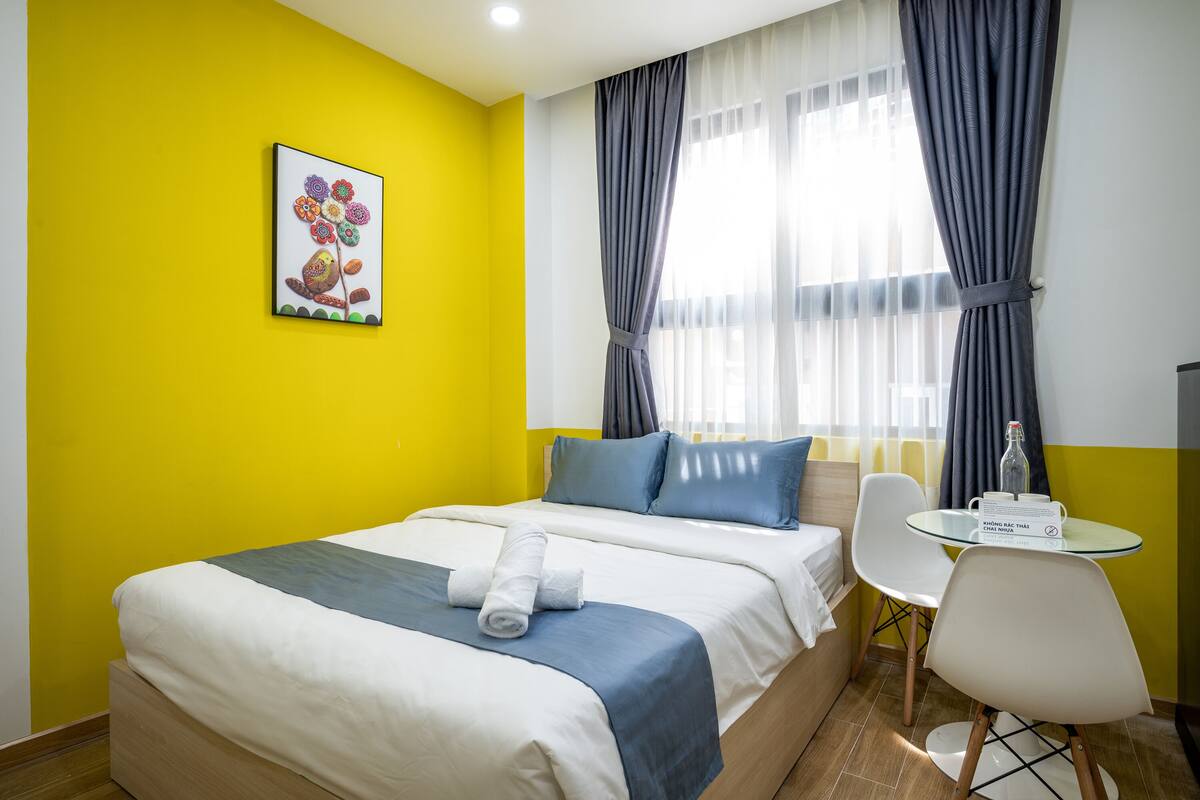 A hotel room with a bed, a small table with a bottle and glasses, a chair, and a painting on the wall.
