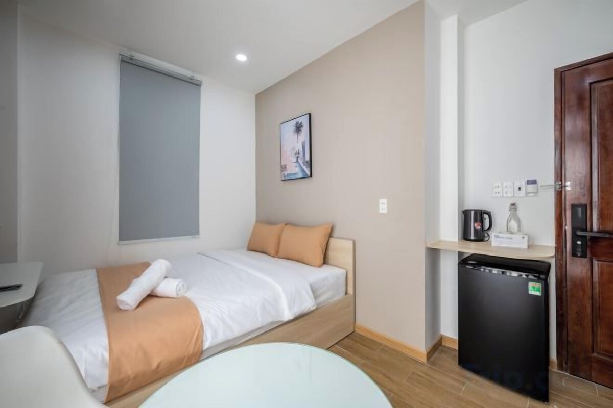 A compact hotel room with a bed, a small desk, a refrigerator, and a door.
