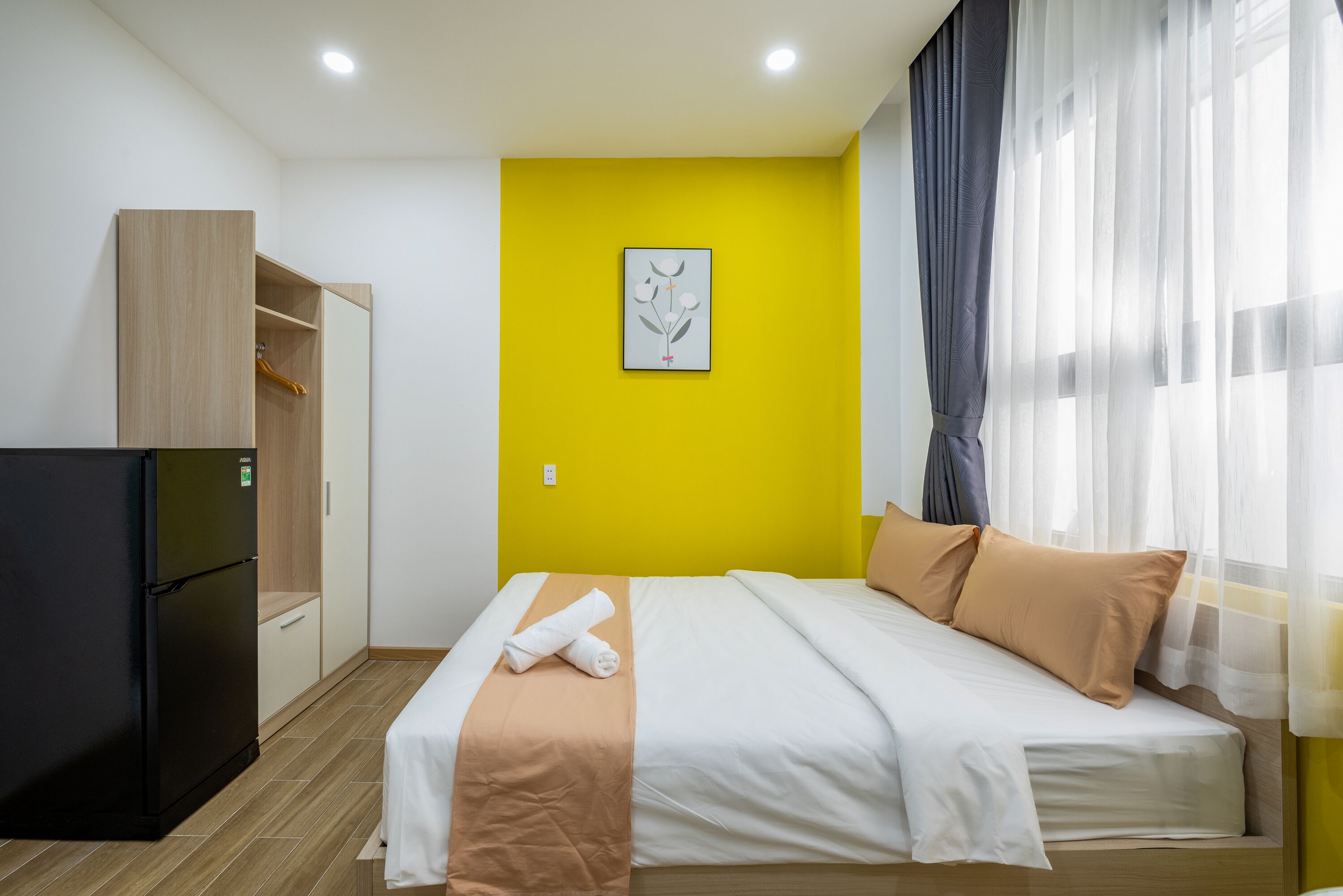 Habitación de hotel con cama, armario, refrigerador negro pequeño y pared de acento amarilla.