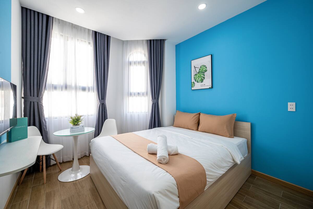 A hotel room with a bed, a desk, a chair, a small table with a plant, and a framed picture on the wall.