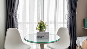 A hotel room with a round glass table, two white chairs, a potted plant, and a bed with a white headboard. - Cozrum Homes - Selina Residence (Ho Chi Minh City)
