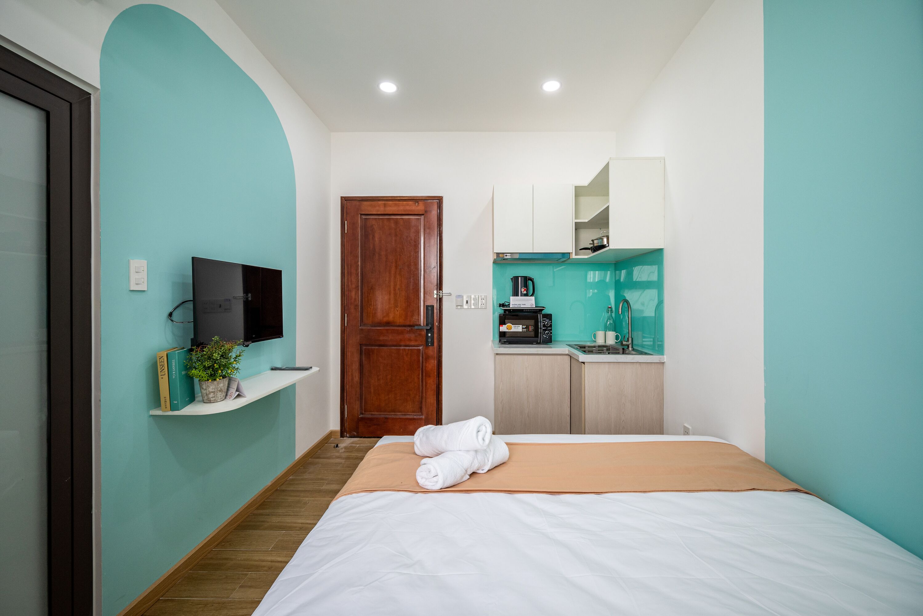 Quarto de hotel pequeno com cama, televisão, cozinha compacta e porta.