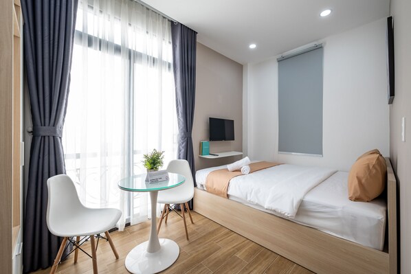 A modern hotel room with a bed, a small table with a chair, a TV, and a window with curtains.
