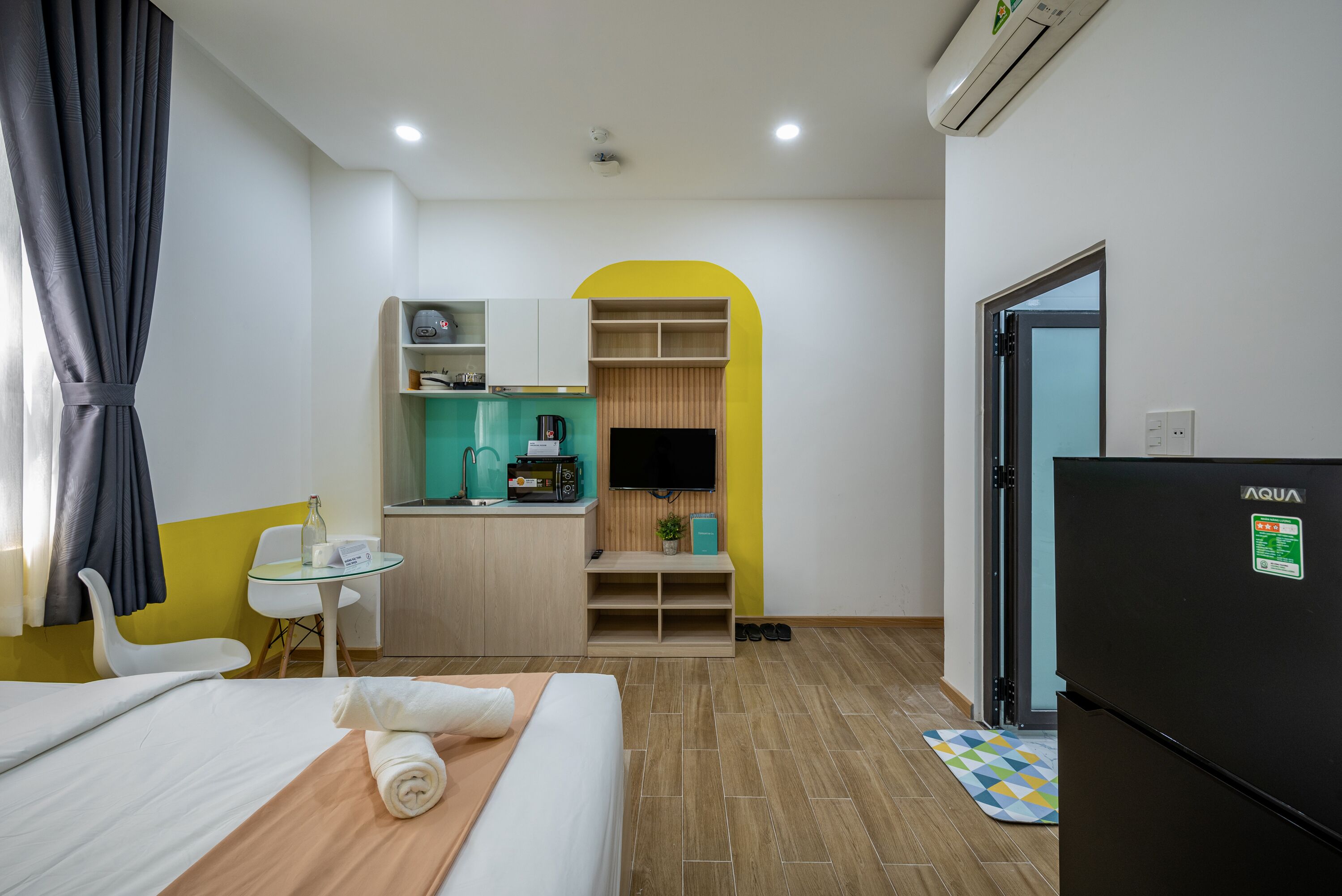 A compact hotel room with a bed, a small kitchenette, a TV, and a refrigerator.