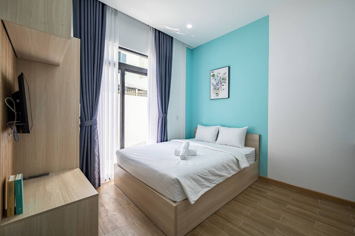 A modern bedroom with a wooden bed, a TV mounted on the wall, and a window with curtains.