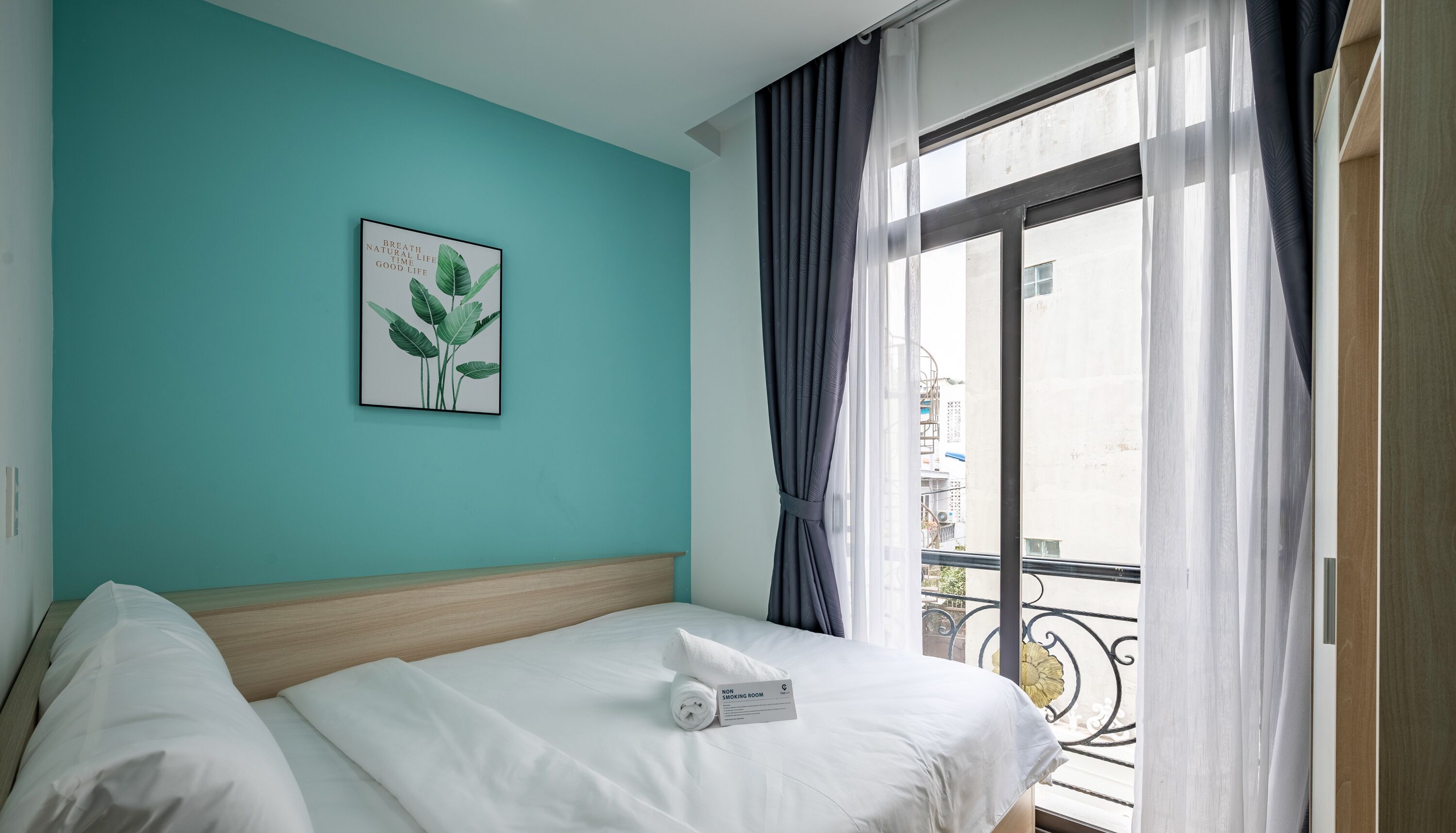 Une chambre d’hôtel avec un lit, une plante en pot et un balcon donnant sur des immeubles.