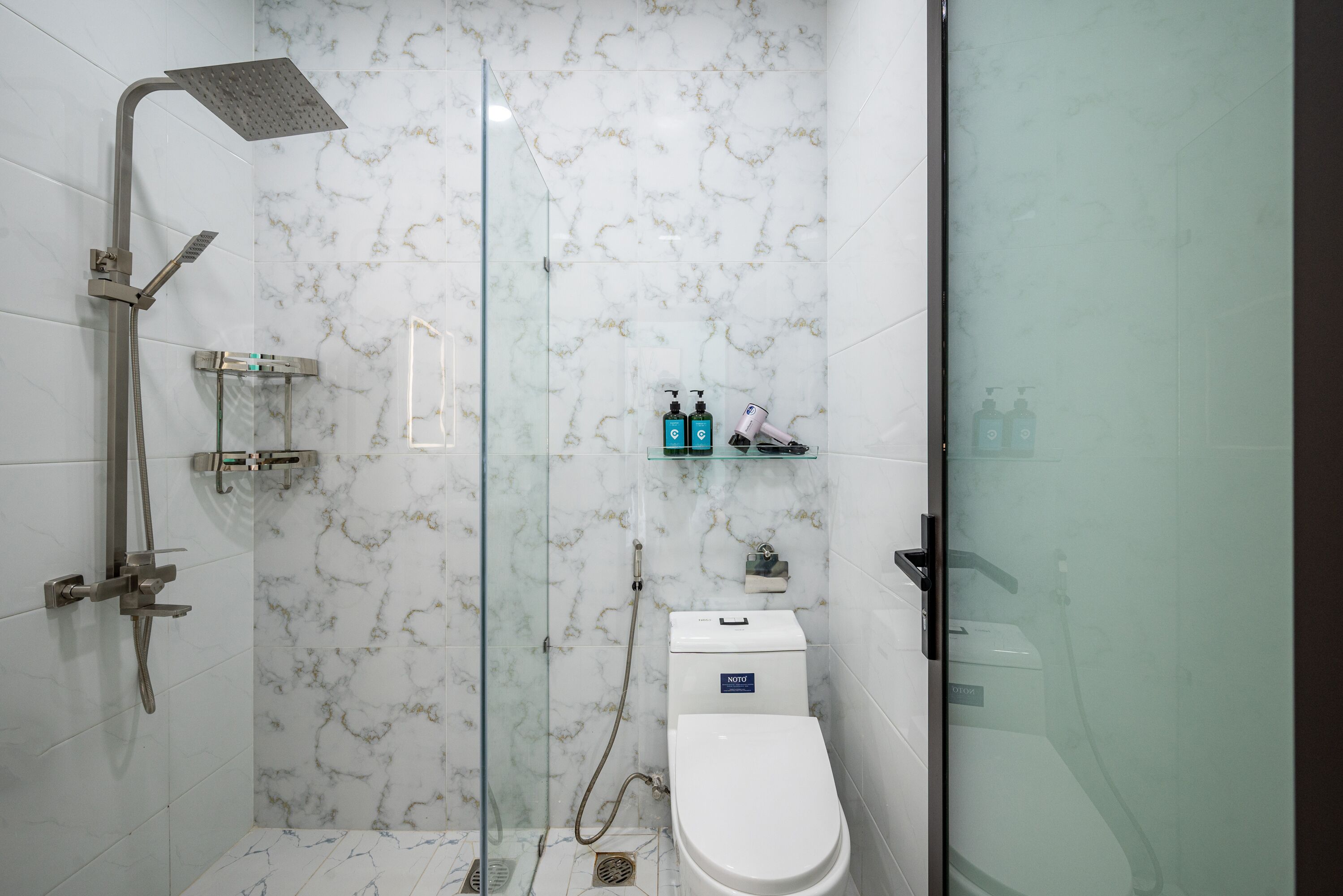 A modern bathroom with a glass shower enclosure, a toilet, and a small shelf with toiletries.