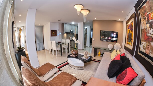Un salon moderne avec un canapé, deux fauteuils, une table basse et un téléviseur. On aperçoit une cuisine à l’arrière-plan, avec des placards et un four à micro-ondes.