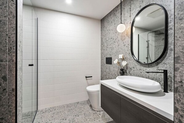 A modern bathroom with a white toilet, a large oval mirror, and a white sink with a dark countertop.