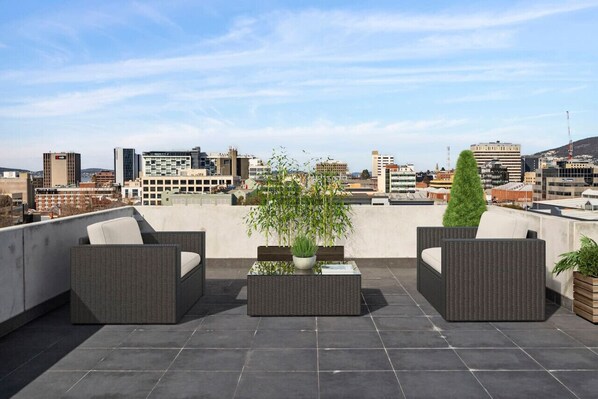 A rooftop terrace with wicker furniture, potted plants, and a cityscape view.
